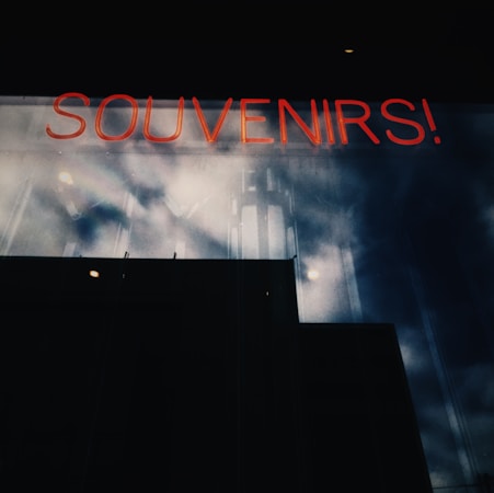 A neon sign reading 'SOUVENIRS!' is visible against a dark reflective glass surface with cloudy skies mirrored on it, creating a moody, industrial atmosphere.