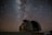 A large, rounded observatory dome sits beneath a vast, star-filled night sky. The Milky Way is prominently visible, with countless stars scattered across the dark backdrop. A faint, linear streak suggests a meteor or satellite passing by.