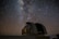 A large, rounded observatory dome sits beneath a vast, star-filled night sky. The Milky Way is prominently visible, with countless stars scattered across the dark backdrop. A faint, linear streak suggests a meteor or satellite passing by.