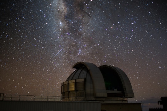 A large, rounded observatory dome sits beneath a vast, star-filled night sky. The Milky Way is prominently visible, with countless stars scattered across the dark backdrop. A faint, linear streak suggests a meteor or satellite passing by.