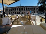 Outdoor café setting with tables and chairs covered in white tablecloths, overlooking an ancient amphitheater. A wine glass and menu are placed on the table, while people walk and cycle along the street under a clear blue sky.