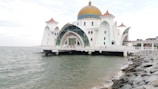 A mosque standing on the water features a large, golden dome framed by angular white walls and arches with intricate designs. The structure is supported by pillars in the sea and is surrounded by a rocky shoreline. Several small domes with reddish-brown tops adorn the corners.