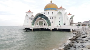 A mosque standing on the water features a large, golden dome framed by angular white walls and arches with intricate designs. The structure is supported by pillars in the sea and is surrounded by a rocky shoreline. Several small domes with reddish-brown tops adorn the corners.