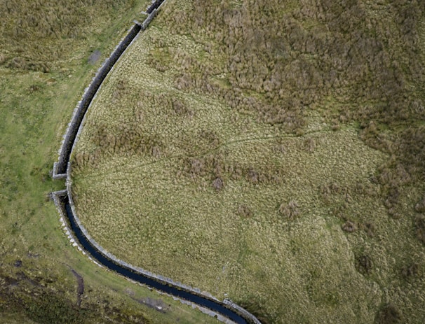 An aerial view of a landscape with a distinct stone wall curving through grassy terrain. The wall appears to separate two sections of the land, with a small waterway running alongside it. The area is covered in patches of brown and green grass, suggesting a natural, undeveloped environment.