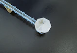 An aerial view of a hexagonal platform, likely a pavilion or gazebo, extending over a body of water. It is connected to the shore by a narrow walkway or bridge painted in light blue, supported by stilts.