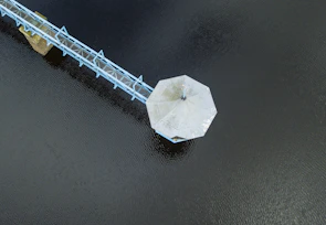 An aerial view of a hexagonal platform, likely a pavilion or gazebo, extending over a body of water. It is connected to the shore by a narrow walkway or bridge painted in light blue, supported by stilts.