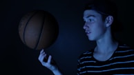 A young person with a focused expression is skillfully balancing a basketball on their finger. They are wearing a striped shirt and a backwards cap. The lighting is dark and moody, creating a dramatic effect with blue hues casting shadows.