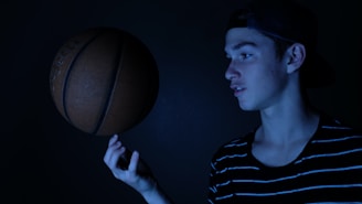 A young person with a focused expression is skillfully balancing a basketball on their finger. They are wearing a striped shirt and a backwards cap. The lighting is dark and moody, creating a dramatic effect with blue hues casting shadows.