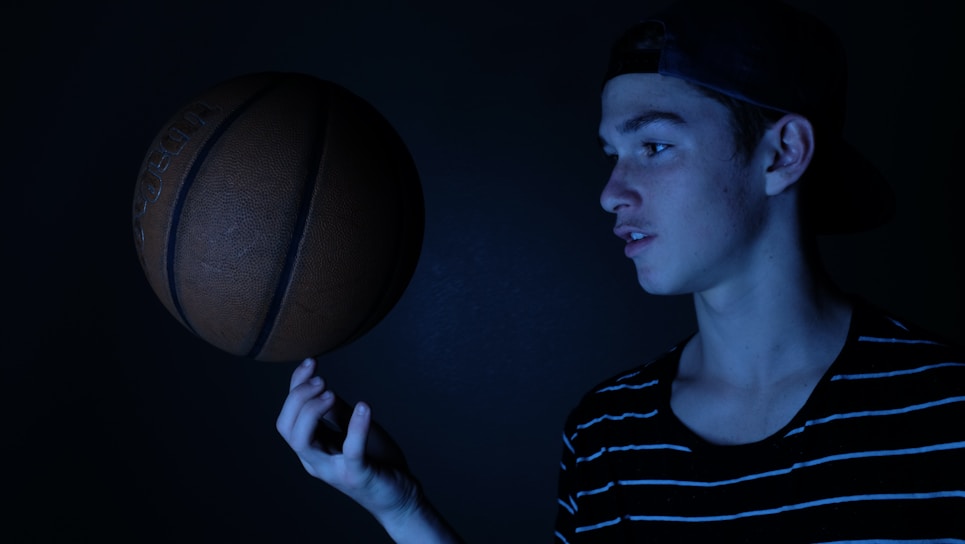 A young person with a focused expression is skillfully balancing a basketball on their finger. They are wearing a striped shirt and a backwards cap. The lighting is dark and moody, creating a dramatic effect with blue hues casting shadows.