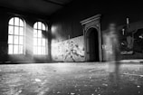 A dimly lit, abandoned room with large arched windows letting in streams of light. Graffiti covers sections of the walls, and there is a ghostly, transparent figure in motion near an ornate doorway. The floor has debris scattered across it, adding to the sense of neglect.