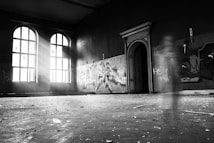 A dimly lit, abandoned room with large arched windows letting in streams of light. Graffiti covers sections of the walls, and there is a ghostly, transparent figure in motion near an ornate doorway. The floor has debris scattered across it, adding to the sense of neglect.
