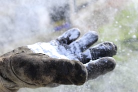 A close-up of a weathered, gloved hand covered in a layer of white powder, possibly flour or dust. The glove appears to be rugged and used for protection, possibly in construction or gardening. The background is blurred with a hint of green and purple tones.