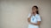 A person in a light blue uniform stands against a plain beige wall, smiling and looking to the side. They have one hand on their chin and the other arm crossed. A lanyard with ID cards is around their neck.