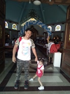 A man and a young girl are standing inside a church. The man is wearing a white shirt with a colorful cartoon design, camouflage pants, and black sneakers. The girl is dressed in a red outfit with white shoes. There are decorated balloons forming an arch in the background. The church features stained glass windows and wooden pews.