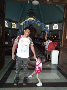 A man and a young girl are standing inside a church. The man is wearing a white shirt with a colorful cartoon design, camouflage pants, and black sneakers. The girl is dressed in a red outfit with white shoes. There are decorated balloons forming an arch in the background. The church features stained glass windows and wooden pews.