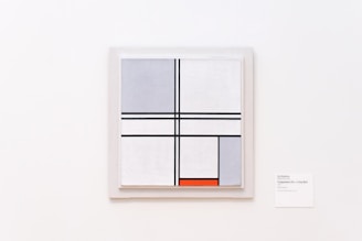 A minimalist abstract painting featuring intersecting black lines forming various geometric shapes against a predominantly white background. Some of the sections are filled with shades of gray, while one small rectangular section at the bottom is colored bright red. The painting is displayed on a plain white wall with a small label next to it.