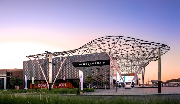 A contemporary shopping center with a unique, modern architectural design. Features include a geometric patterned facade and an intricately designed metal canopy. The branding of 'Le BHV Marais' is prominently displayed on the building. Lush greenery surrounds the area, with banners and signs visible, and pavement marks indicating traffic directions.