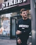 man in black Givenchy sweatshirt standing beside of post in front of Strek store during daytime shallow focus photography