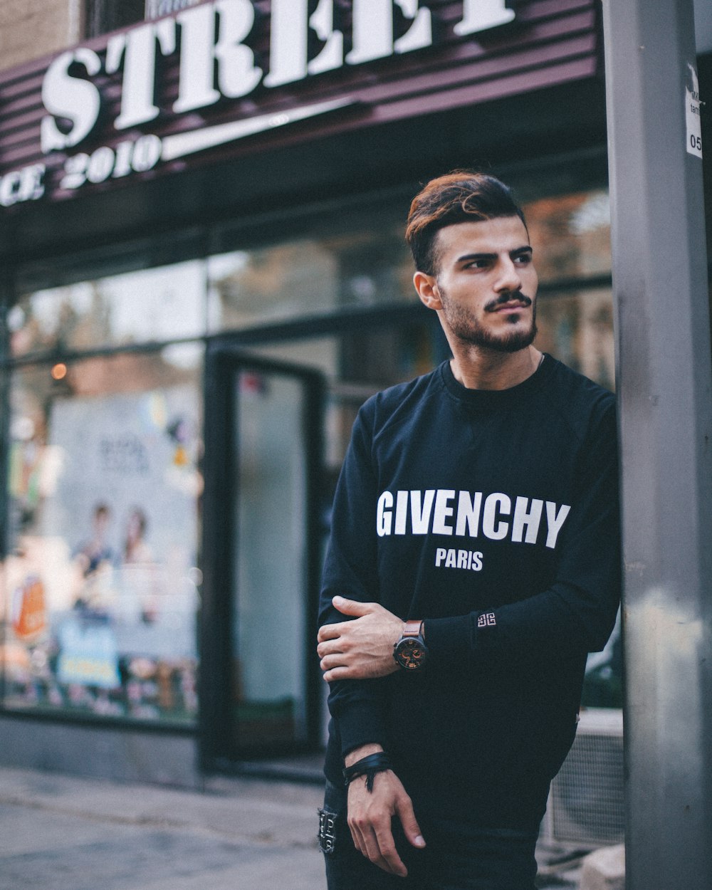 Mann im schwarzen Givenchy-Sweatshirt steht neben dem Pfosten vor dem Strek-Geschäft während der Tagesfotografie mit flachem Fokus
