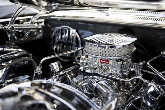 A close-up view of a polished, chrome engine with intricate mechanical components and an air filter marked with the brand 'Edelbrock'. The shiny metal surfaces reflect light, creating a glossy and detailed appearance.