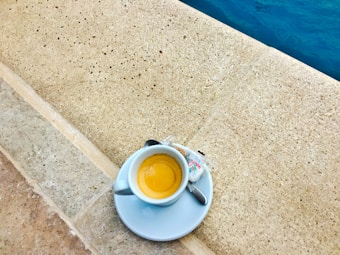 A small cup of espresso is placed on a light stone surface next to a swimming pool. The cup sits on a saucer with a small spoon and a packet of sugar beside it.