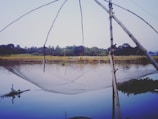 A large fishing net is suspended above a calm body of water, supported by bamboo poles. In the background, there is lush greenery and trees lining the horizon. The water reflects the objects and greenery around it, creating a serene and tranquil scene.