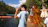 A person is wearing a light blue shirt labeled 'Staff' and is holding a camera while standing by a lake. The location seems to be a recreational area with lush green trees and a wooden cabin nearby. Life vests are hanging on a tree branch, and another person appears to be swimming in the water.