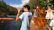A person is wearing a light blue shirt labeled 'Staff' and is holding a camera while standing by a lake. The location seems to be a recreational area with lush green trees and a wooden cabin nearby. Life vests are hanging on a tree branch, and another person appears to be swimming in the water.