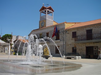 A picturesque town square features a stone building with a clock tower and red tiled roof. A fountain is active in the center, its water jets creating an arc pattern. Flags hang from the building, which is constructed in a traditional style with wrought iron balconies. The area is bathed in sunlight, and there is a clear blue sky overhead.