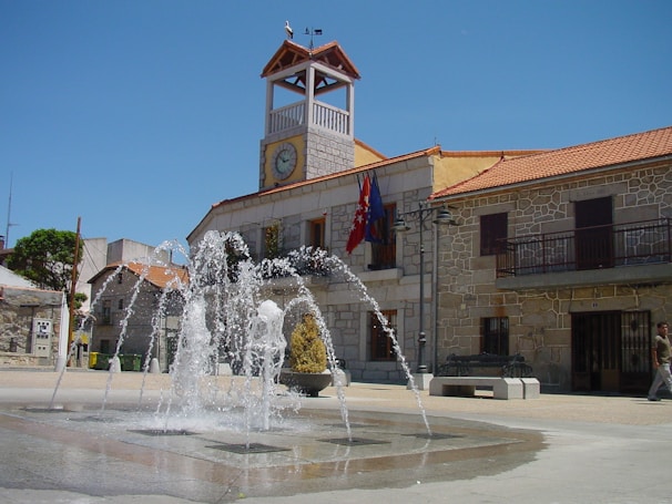 A picturesque town square features a stone building with a clock tower and red tiled roof. A fountain is active in the center, its water jets creating an arc pattern. Flags hang from the building, which is constructed in a traditional style with wrought iron balconies. The area is bathed in sunlight, and there is a clear blue sky overhead.
