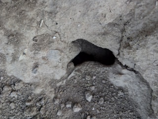 A close-up view of a rough, textured concrete surface with a significant hollow or cavity in the center surrounded by various small gravel particles and irregularities.