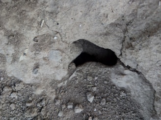 A close-up view of a rough, textured concrete surface with a significant hollow or cavity in the center surrounded by various small gravel particles and irregularities.