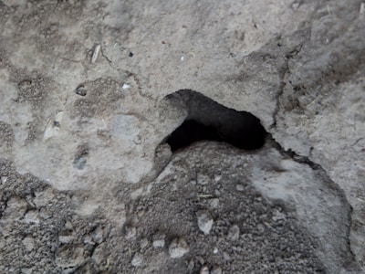 A close-up view of a rough, textured concrete surface with a significant hollow or cavity in the center surrounded by various small gravel particles and irregularities.