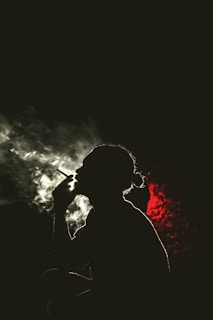 A silhouette of a person smoking with smoke swirling around, set against a dramatic black background and a deep red light illuminating part of the scene.