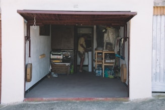An open garage with various objects stored inside, including boxes, bottles, and shelves filled with items. A person is standing inside the garage near the back wall, partially obscured by the shadows.