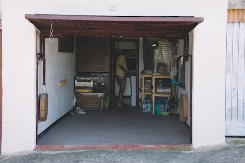 An open garage with various objects stored inside, including boxes, bottles, and shelves filled with items. A person is standing inside the garage near the back wall, partially obscured by the shadows.