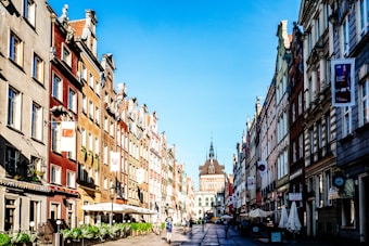 A vibrant city street features colorful, historic buildings lining the road with intricate facades and multiple windows. The street is bustling with people walking and enjoying the cafes that sit beneath canopies. In the distance, a grand building with a spire stands prominent against the clear blue sky.