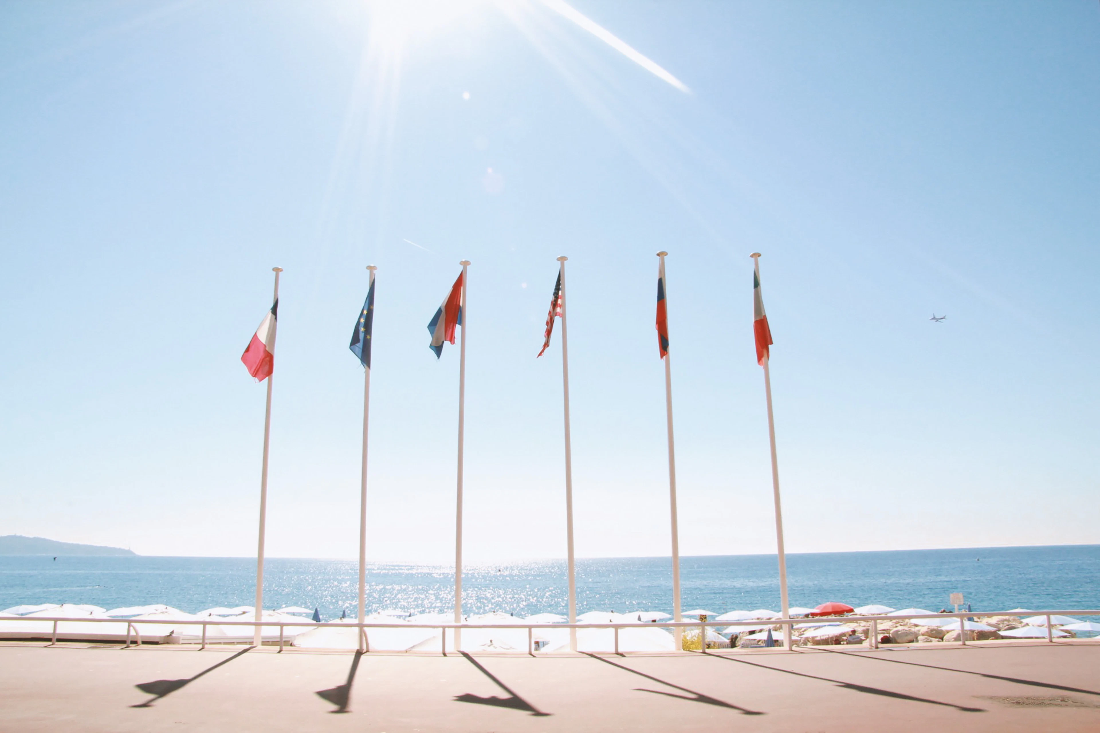 Flags fly along Nice's seafront promenade against a sparkling blue sea and bright sun.