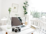 A minimalist workspace featuring a laptop on a white table along with a vintage camera and a vase with white flowers. Natural light filters through large windows, creating a bright and airy atmosphere. A drink with a straw sits next to a notebook and pen. Indoor plants add a touch of greenery in the background.