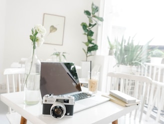 A minimalist workspace featuring a laptop on a white table along with a vintage camera and a vase with white flowers. Natural light filters through large windows, creating a bright and airy atmosphere. A drink with a straw sits next to a notebook and pen. Indoor plants add a touch of greenery in the background.
