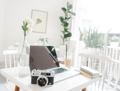 A minimalist workspace featuring a laptop on a white table along with a vintage camera and a vase with white flowers. Natural light filters through large windows, creating a bright and airy atmosphere. A drink with a straw sits next to a notebook and pen. Indoor plants add a touch of greenery in the background.
