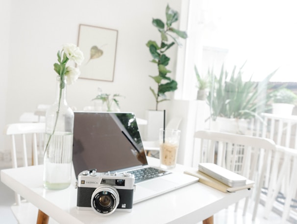 A minimalist workspace featuring a laptop on a white table along with a vintage camera and a vase with white flowers. Natural light filters through large windows, creating a bright and airy atmosphere. A drink with a straw sits next to a notebook and pen. Indoor plants add a touch of greenery in the background.