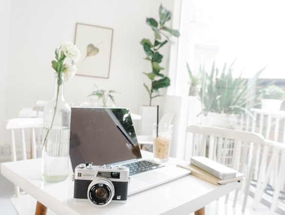 A minimalist workspace featuring a laptop on a white table along with a vintage camera and a vase with white flowers. Natural light filters through large windows, creating a bright and airy atmosphere. A drink with a straw sits next to a notebook and pen. Indoor plants add a touch of greenery in the background.