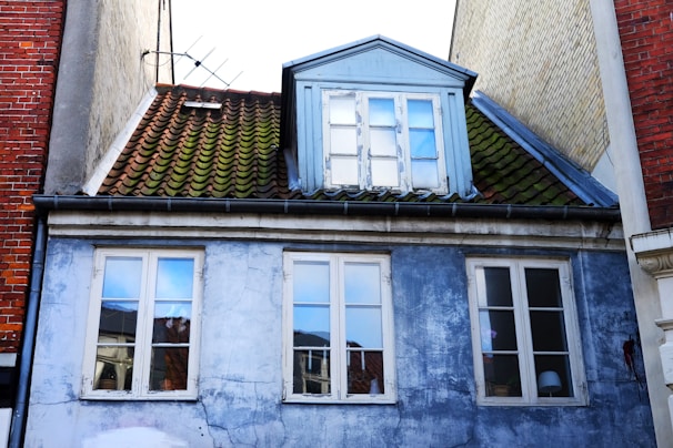 A worn building façade featuring a blue peeling paint surface with multiple windows. The roof consists of green weathered tiles and includes a dormer with a blue frame. The building appears to be situated tightly between other brick structures.