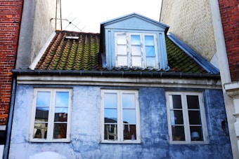 A worn building façade featuring a blue peeling paint surface with multiple windows. The roof consists of green weathered tiles and includes a dormer with a blue frame. The building appears to be situated tightly between other brick structures.