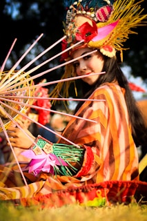 A person in traditional attire holding a colorful, intricate handcrafted object. The costume includes vibrant striped patterns and various accessories. A decorated headpiece with feathers, flowers, and other ornamental details is worn. The background is slightly blurred, suggesting an outdoor event.