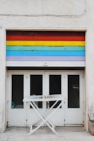 A garage door painted with horizontal rainbow stripes spans above four white double doors with glass panes. In front, there is a white folding drying rack with a few colorful clothespins clipped to it.