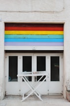 A garage door painted with horizontal rainbow stripes spans above four white double doors with glass panes. In front, there is a white folding drying rack with a few colorful clothespins clipped to it.