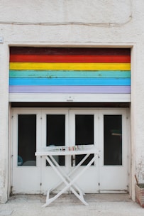 A garage door painted with horizontal rainbow stripes spans above four white double doors with glass panes. In front, there is a white folding drying rack with a few colorful clothespins clipped to it.