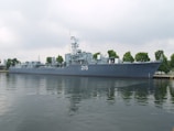A large naval ship with the number 215 painted on its side is moored along a waterfront. The vessel is equipped with various antennas and radar equipment, and several trees are visible in the background, indicating a dock or port area. The water reflects the image of the ship, creating a calm and orderly scene under a cloudy sky.
